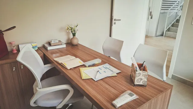 A mesa de madeira do consultório, com cadeiras brancas, flores e a porta aberta para o corredor